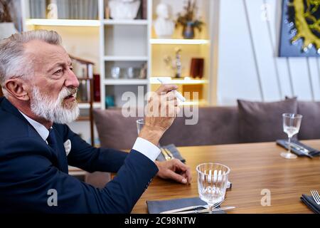 Senior grey haired smoker man keep in hands smoke free-cigarette, explore cigarette, male sits in restaurant Stock Photo