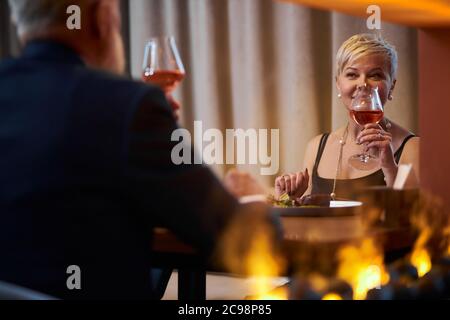 Happy gray-haired elderly lady in black dress toasting with glass of champagne to male in elegant restaurant, look at him. Rear view on man in tuxedo. Stock Photo