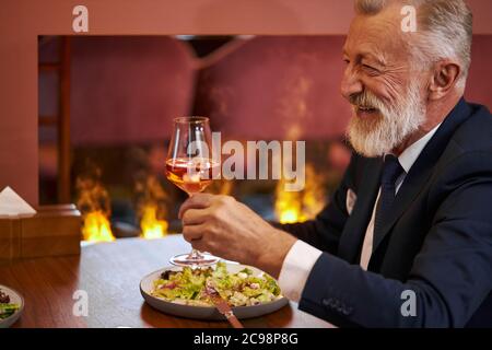 Senior elegant bearded and gray-haired man in tuxedo with glass of champagne sit in restaurant and laugh, look at person opposite. Background fireplac Stock Photo