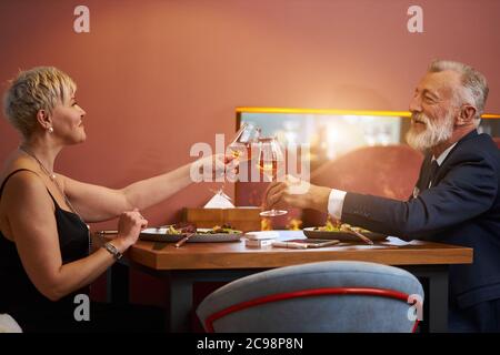 Senior elegant couple sit in restaurant and clink glasses with champagne, celebrate. Bearded gray-haired man in tuxedo and caucasian woman in black dr Stock Photo