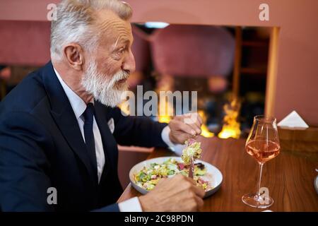Elegant caucasian senior male sits in restaurant. Bearded man look and eat. Fireplace behind him. Horizontal image Stock Photo
