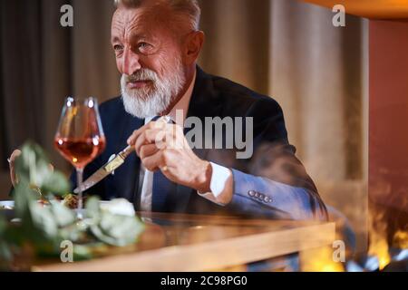 Mature beared businessman in tidy beautiful suit eat and drink champagne in romantic atmosphere. Light smoke from the fireplace on the side. Restauran Stock Photo