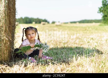beautiful little girl with a bouquet of field daisies sits in the shade of a haystack Stock Photo