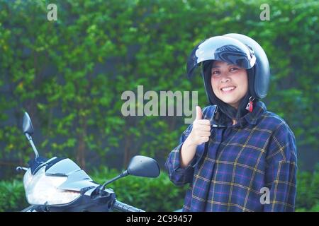 A Young Woman fastening or Wearing her motorbike helmet with blur Fukien tea tree Carmona retusa green background - Zero accident concept Stock Photo