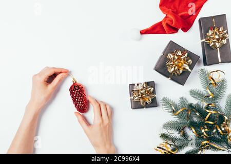 Female's hands holding red Christmas tree toy fir cone next to pine branches, gift boxes, golden ribbons and Santa Claus hat on white table, flat lay. Stock Photo