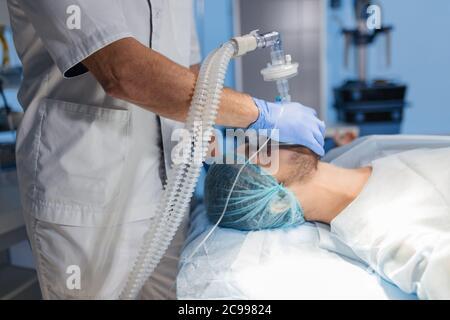 Male assistants prepares a patient to invasive surgery in the Hospital operating room, holding oxygen mask over man s face, close up Stock Photo