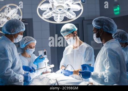 Urgent surgery. Professional smart intelligent surgeons standing near the patient and performing an operation while saving his life. Professional trea Stock Photo