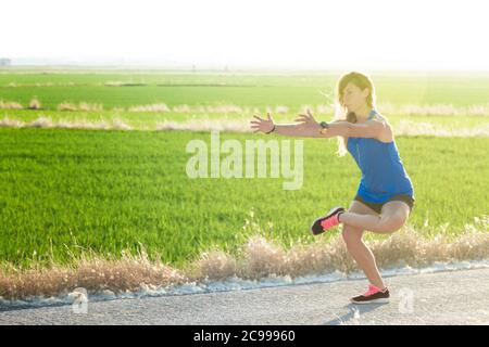 A young athlete girl in blue t-shirt doing stretches on the road next to a rice field at sunset Stock Photo