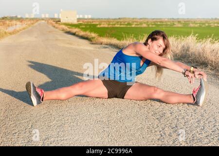 A young athlete girl in blue t-shirt sitting doing leg stretches on the road next to a rice field at sunset Stock Photo