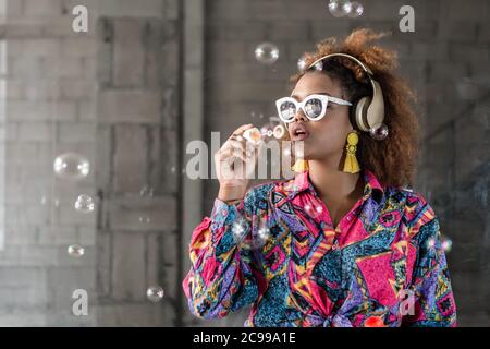 Trendy African american woman in wireless headset and bright earrings standing with open mouth while blowing bubbles and having fun near brick wall Stock Photo