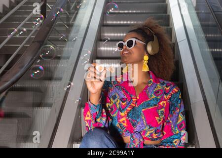 Low angle of cheerful female in bright wear and bijouterie blowing fragile and transparent bubbles while sitting on moving stairs in wireless headset Stock Photo