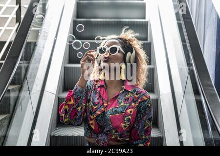 Trendy anonymous black lady in yellow earrings and sunglasses blowing bubbles while sitting with pout lips on moving staircase in wireless headset in Stock Photo