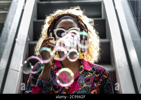 Anonymous African American female in headphones and colorful blouse blowing transparent bubbles while sitting on moving staircase in back lit Stock Photo