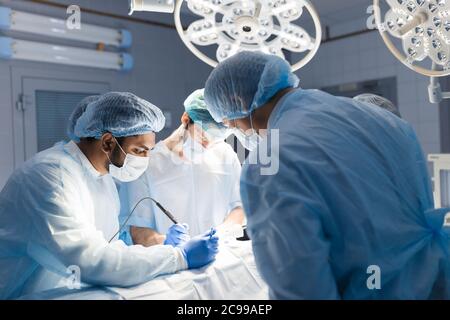 Surgeon doctor operating using electric scalpel wearing blue surgical mask and surgical cap in surgery room with his team surgeons operating Stock Photo