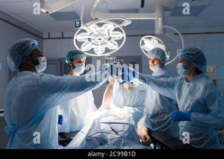 Multiethnic surgeons team joining hands together with glasses of apple juice, cheering after rescue patient in operation room at hospital. Stock Photo