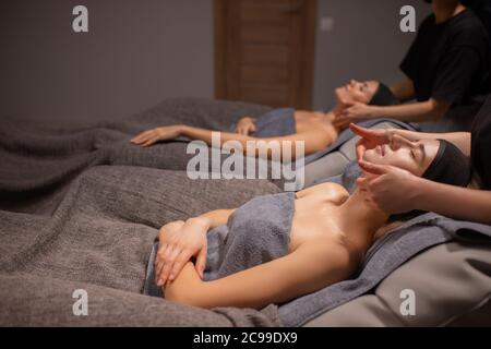 two beautiful caucasian women came in spa to get face lift procedure, they lie on bed and relax while getting massage on face and head by professional Stock Photo