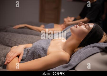 side view on calm relaxed caucasian woman lying in wellbeing salon before skin treatment, woman get face lift massage in the background Stock Photo