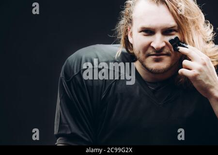 Young caucasian blonde man in american football player's ammunition wearing black painted stripes on his cheeks looking at camera against black backgr Stock Photo