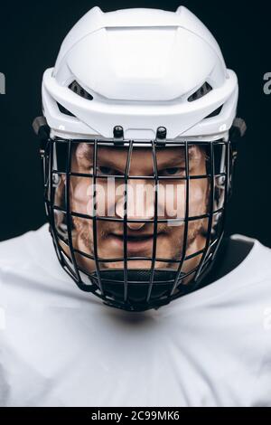 Portrait of hockey keeper in protective helmet with carbonic defence in white uniform grinning at camera over black background Stock Photo