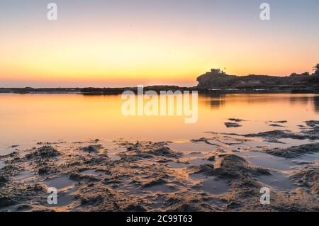 Sunset long exposure shot showing a calm sea lake lagoon with rocks in the foreground and a temple on an overlooking hill in the distance Stock Photo