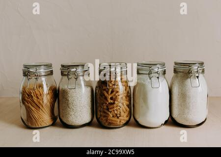 Five glass jar containers full with dried uncooked food ingredients. Rice, pasta, flour, sugar. Stock Photo