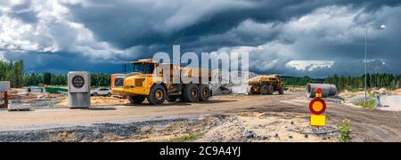 Panorama view on two heavy industrial yellow dump trucks parked close to construction site with stocked building materials, dark blue clouds before ra Stock Photo