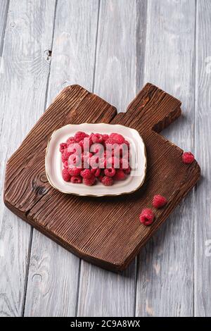 Raspberry fruits in plate on old teak cutting board, healthy pile of summer berries on wooden background, angle view Stock Photo