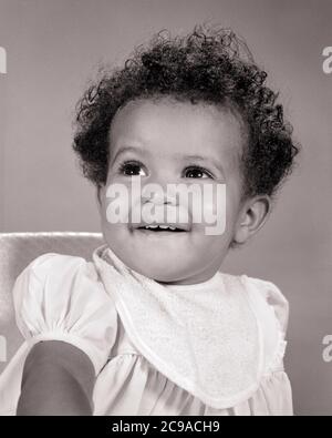 1960s ANTICIPATION SMILING HUNGRY AFRICAN-AMERICAN BABY GIRL SITTING IN HIGHCHAIR WEARING BIB WAITING TO BE FED - n1969 HAR001 HARS AFRICAN-AMERICAN BE LOW ANGLE PRIDE IN TO ANTICIPATION SMILES CONCEPTUAL JOYFUL STYLISH BIB GROWTH BABY GIRL BLACK AND WHITE HAR001 OLD FASHIONED Stock Photo