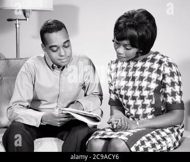 1960s AFRICAN-AMERICAN COUPLE SITTING ON COUCH HUSBAND SHOWING WIFE ARTICLE IN NEWSPAPER SHE IS WEARING DRESS HOUNDSTOOTH PRINT - n2234 HAR001 HARS LIFESTYLE FEMALES MARRIED STUDIO SHOT SHOWING SPOUSE HUSBANDS HEALTHINESS HOME LIFE COPY SPACE HALF-LENGTH LADIES PERSONS INSPIRATION CARING MALES B&W PARTNER SUCCESS DREAMS HAPPINESS AFRICAN-AMERICAN SHE PRIDE CONCEPTUAL STYLISH PERSONAL ATTACHMENT AFFECTION EMOTION HOUNDSTOOTH MID-ADULT MID-ADULT MAN MID-ADULT WOMAN TOGETHERNESS WIVES BLACK AND WHITE HAR001 OLD FASHIONED Stock Photo