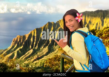 Hawaii hiker girl tourist doing shaka hawaiian hand sign at Na pali lookout in Kauai, Hawaii. Travel Asian woman backpacker hiking with bag at famous Stock Photo