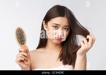 Upset stressed young Asian woman holding damaged dry hair on hands over white isolated background. Stock Photo