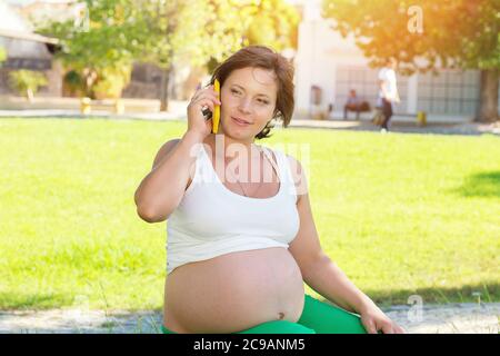 Happy pregnant woman talking on phone at sunset in a park sitting outdoors on a green grass meadow in a city park on a sunny day Stock Photo