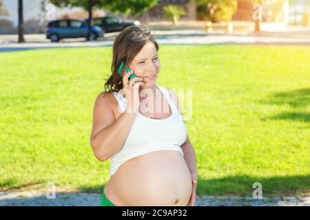 Happy pregnant woman talking on phone sitting at sunset in a park outdoors on a green grass meadow Stock Photo