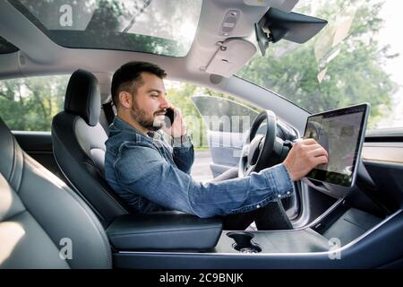 Testing a new electric futuristic car with self driving system. Side view of satisfied Caucasian man in casual jeans shirt sitting in modern car Stock Photo