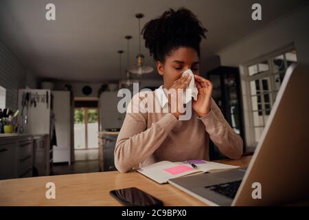 Sick exhausted young woman suffering from cold virus and coughing using paper napkins while working from home