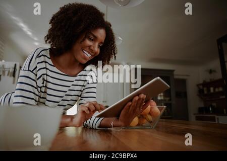 Happy woman leaning on kitchen counter watching cooking videos in digital tablet in kitchen Stock Photo