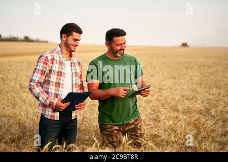 Two farmers stand in wheat stubble field, discuss harvest, crops. Senior agronomist with touch tablet pc teaches young coworker. Innovative tech