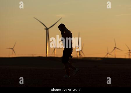 A man walks talking on the phone in the countryside at sunset, after taking pictures of distant wind turbines, on the horizon. Stock Photo