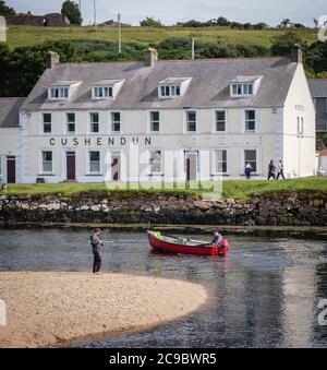 Man fishing from the shore as red boat goes out to sea from Cushendun in County Antrim, Northern Ireland passing the Cushendun Hotel Stock Photo