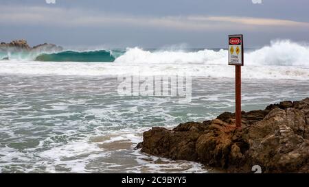 A warning sign with a big wave breaking selectively blurred on the beach at Petrel Cove located on the Fleurieu Peninsula Victor Harbor South Australi Stock Photo