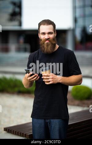Young man with beard holding coffee cup and talking on mobile phone while walking outdoors. Stock Photo