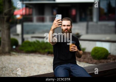 Young bearded brutal man have video call converation sitting on the bench on street Stock Photo