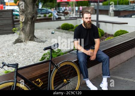 Coffee on the go. Side view of young bearded man drinking coffee while sitting on his bicycle outdoors Stock Photo