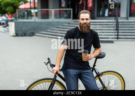 Coffee on the go. Side view of young bearded man drinking coffee while sitting on his bicycle outdoors Stock Photo