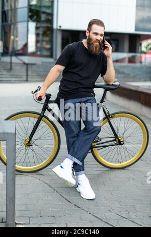 Happy young bearded man talking on the mobile phone and smiling while sitting near his bicycle outdoors Stock Photo