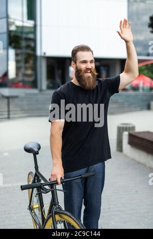 Young bearded man with fixed gear bike waving hand on city street Stock Photo