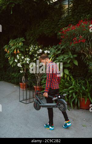 A handsome young bearded caucasian man with sunglasses is standing on the street with e scooter Stock Photo