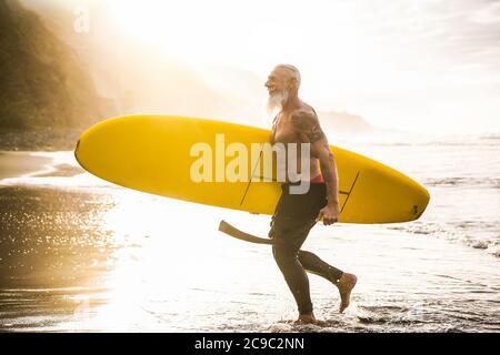 Senior fit guy walking with longboard after surfing at sunset - Mature tattooed man having fun doing extreme sport on tropical beach - Joyful elderly Stock Photo
