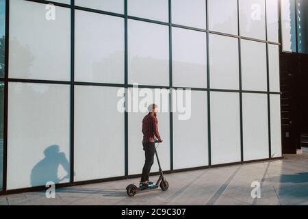 A handsome young bearded businessman rides e scooter in front of the white wall of the building Stock Photo