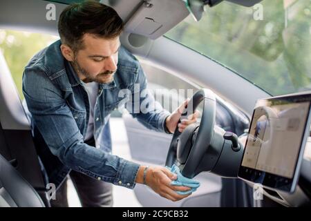 Cleaning of car interior, car detailing concept. Young Caucasian man in casual wear washes a car interior, car steering wheel with blue microfiber Stock Photo